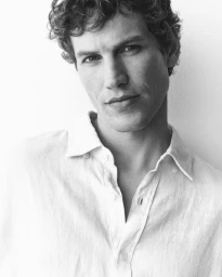 Black and white portrait of Paul Hupfer wearing an open white linen shirt against a bright seamless background.