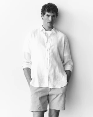 Model Paul Hupfer wearing a white linen button-down shirt and shorts standing against a white studio background.