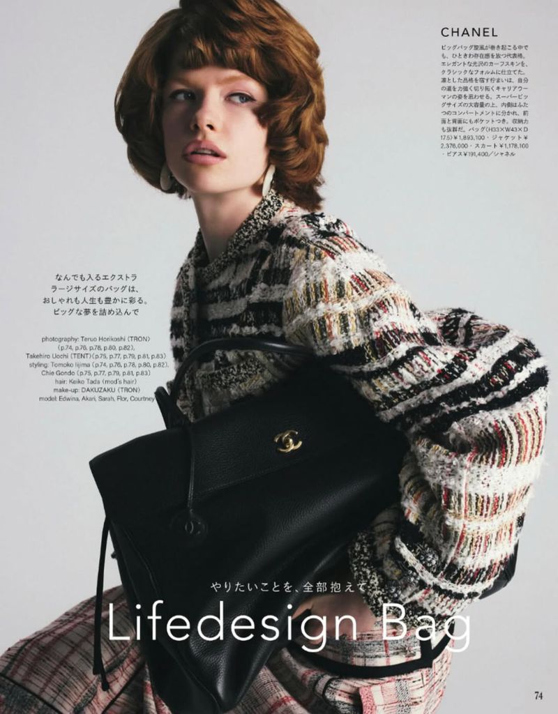 Model with reddish-brown hair wearing a tweed jacket and holding a large black leather handbag in a studio.