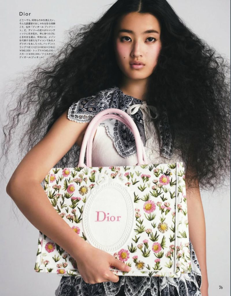 East Asian model with voluminous dark wavy hair holding a floral embroidered Dior tote bag with pink handles against a pale background.
