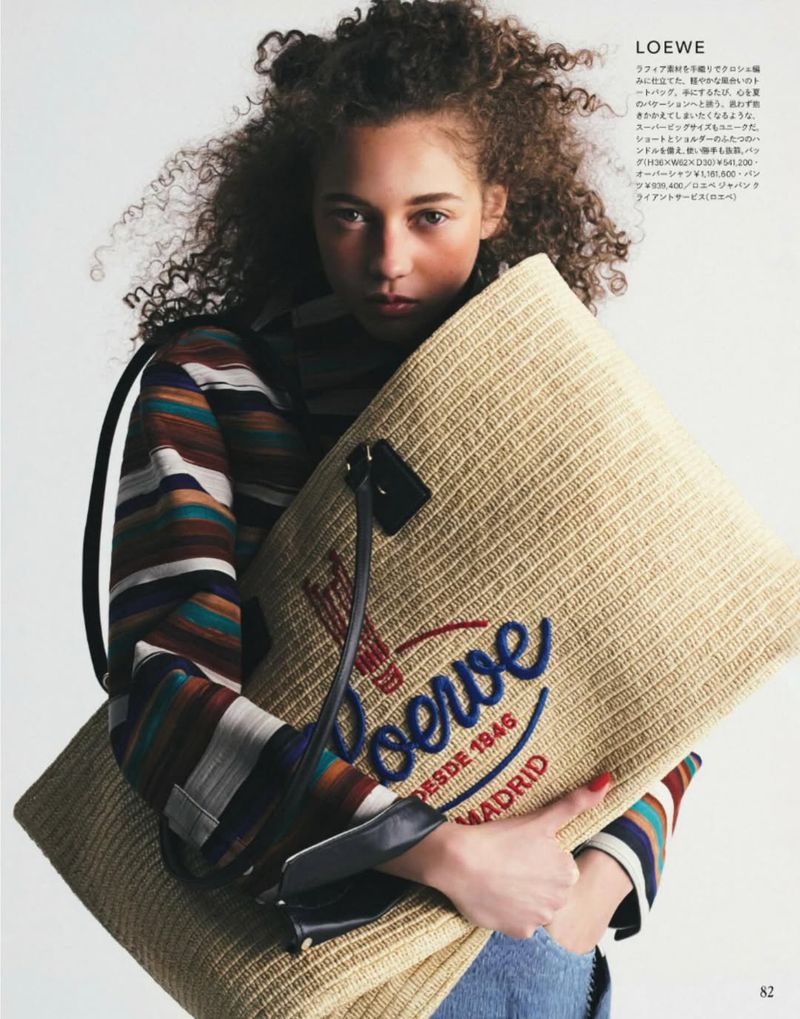 A curly-haired model wearing a striped shirt holds a large woven straw tote bag in front of a white studio backdrop.