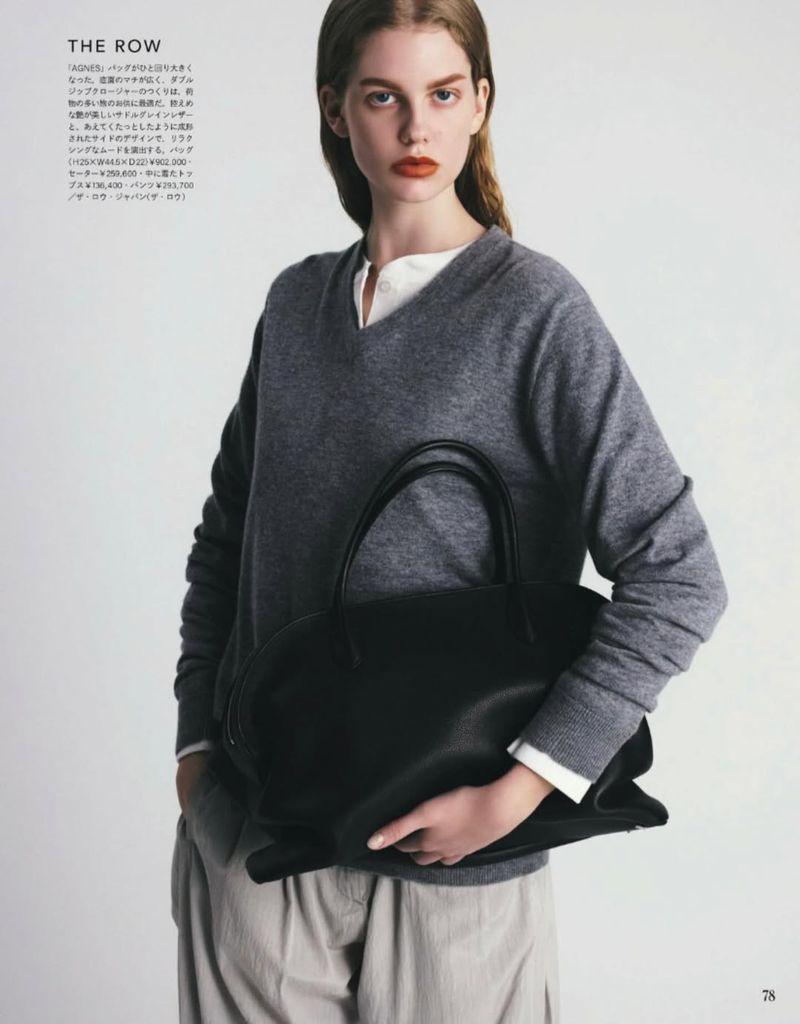 Model wearing a grey v-neck sweater and beige trousers holding a large black leather handbag against a white studio background.