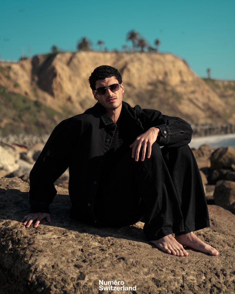 Man in sunglasses and an oversized black jacket sitting barefoot on coastal rocks before a sunlit cliff.