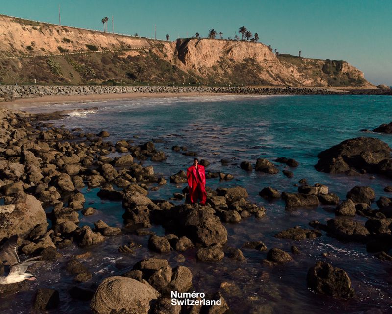 Model wearing a vibrant red coat standing on a rocky ocean shore beneath coastal cliffs.