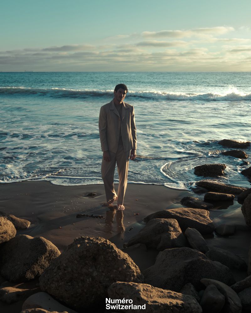 A man in a beige tailored suit stands barefoot on wet sand along a rocky beach shoreline with ocean waves rolling behind him.
