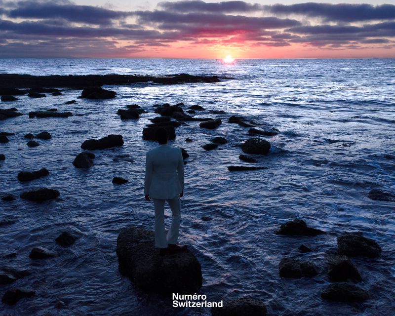 Man in a light suit standing on a rocky shoreline looking out at a vibrant sunset over the ocean.