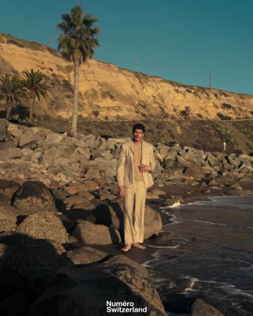 Model in a tailored beige suit stands barefoot on a rocky shoreline beneath a sunlit coastal cliff.