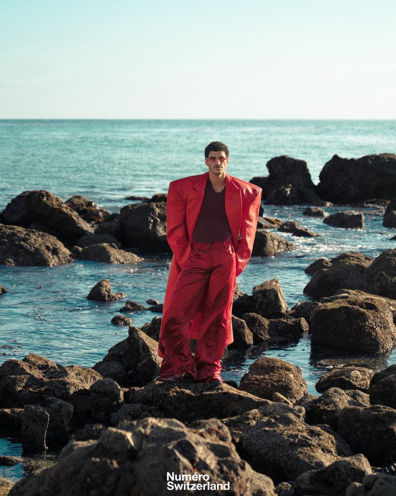 Man wearing a structured red coat with exaggerated shoulders standing on dark coastal rocks with ocean waves in the background.