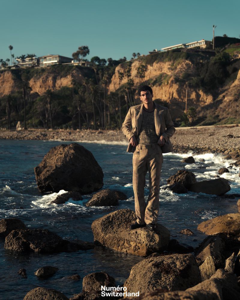 A man wearing a tailored beige suit stands posed on coastal rocks by crashing waves with hills in the background.