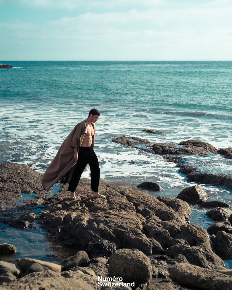 Barefoot man in an open taupe robe coat and black trousers walking across a rocky shoreline by ocean waves.