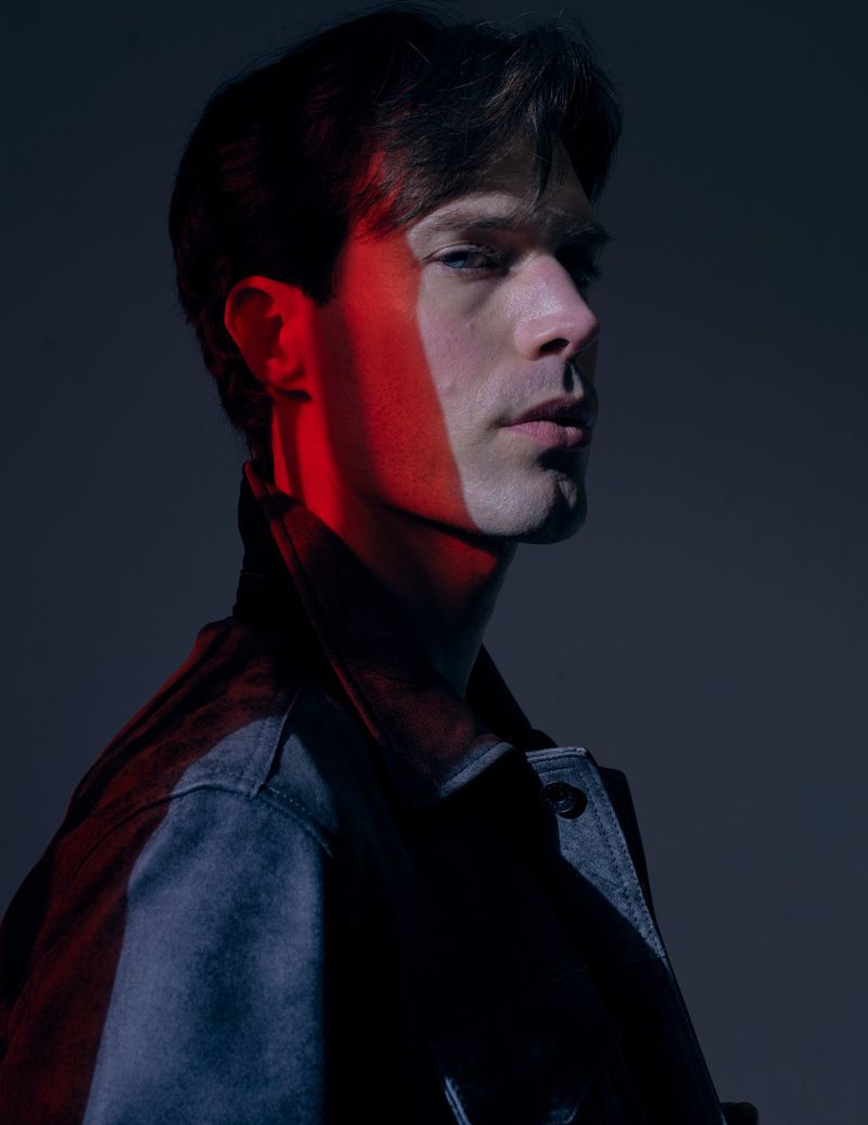 Close-up portrait of a man in profile bathed in a sharp red beam of light and cool shadows against a dark background.
