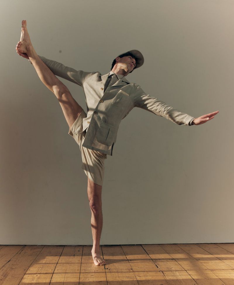Male dancer balancing on one leg in a beige tailored jacket and shorts against a neutral studio wall with wooden floors.
