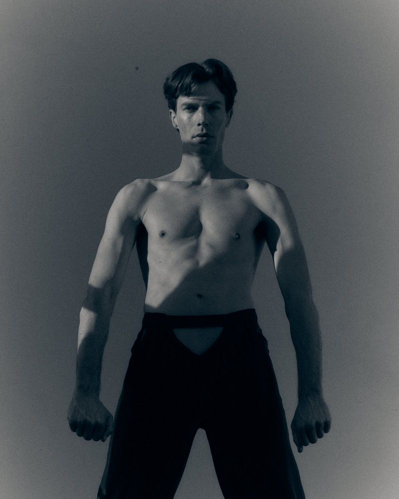 Shirtless man standing against a grey studio backdrop wearing black cutout trousers with sharp shadows across his torso.