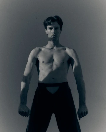 Shirtless man standing against a grey studio backdrop wearing black cutout trousers with sharp shadows across his torso.