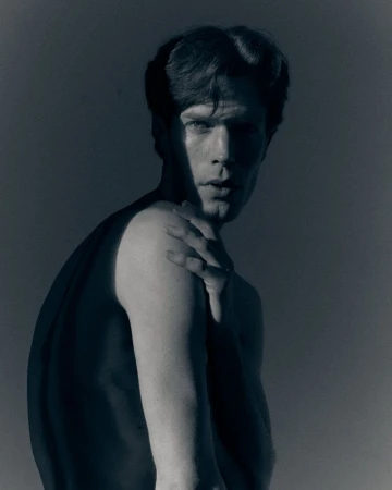 Man posing with hand on shoulder under dramatic chiaroscuro lighting against a muted backdrop for Essay on Oneself editorial.
