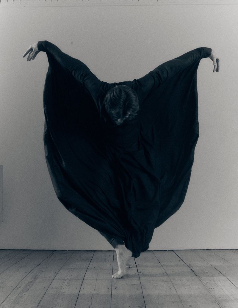 Dancer in a flowing black draped garment posed barefoot on a wooden floor against a plain wall.