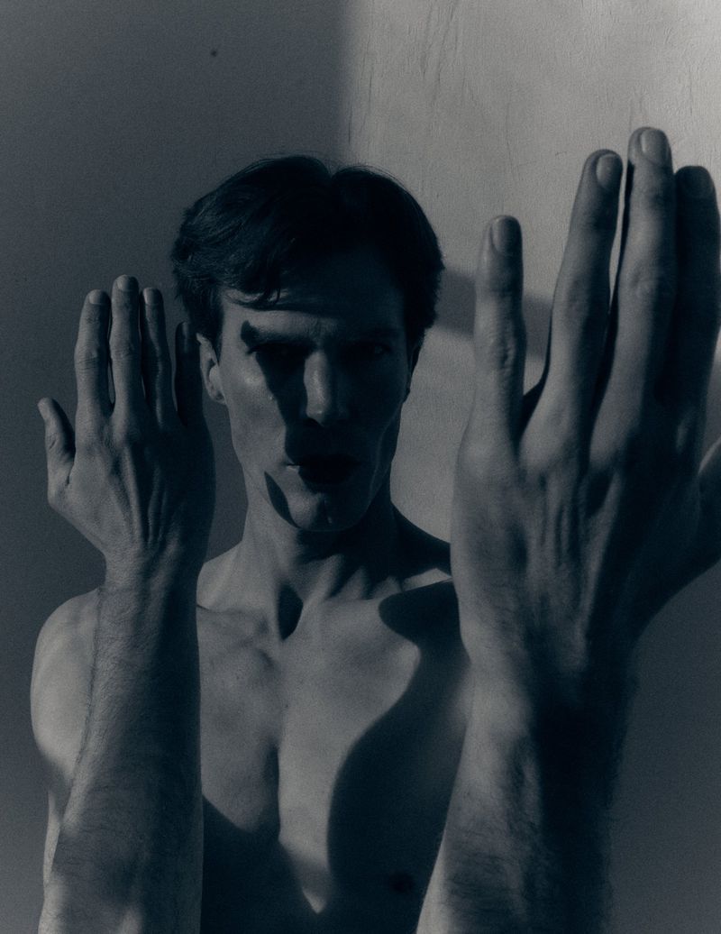 Shirtless man with raised hands creating dramatic shadows across his face against a corner wall.