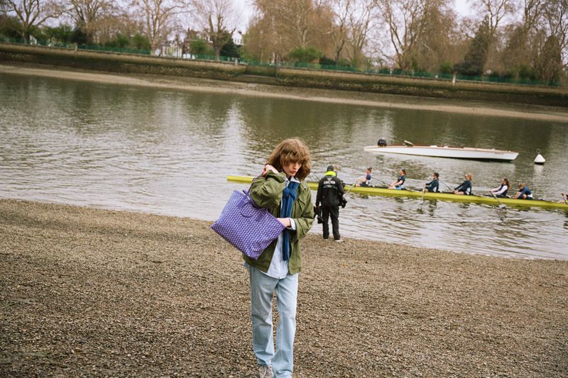 Model carrying a purple patterned Moynat tote bag along a pebbled riverbank with rowers practicing on the water.