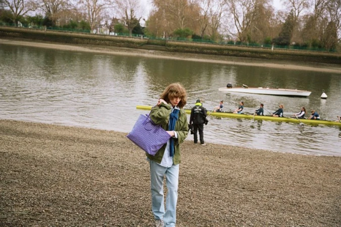 Model carrying a purple patterned Moynat tote bag along a pebbled riverbank with rowers practicing on the water.