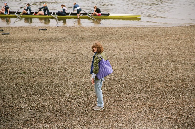 Model Katherine Vinten holding a purple tote bag on a pebbled shoreline with rowers in a boat on the water behind her.