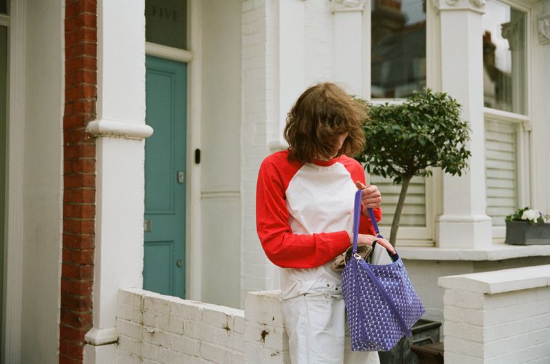 Model Katherine Vinten in a red and white raglan shirt looking into a purple monogram tote bag in front of a townhouse.