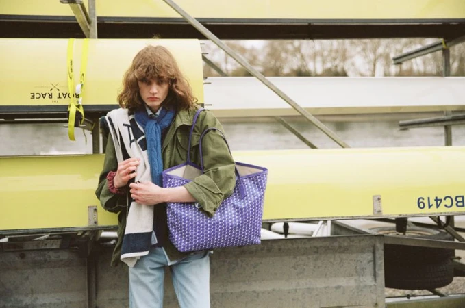Model Katherine Vinten holding a purple patterned tote bag while wearing an olive jacket beside stacked yellow rowing boats.