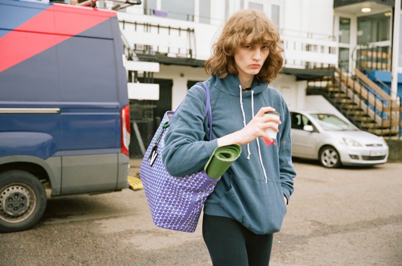 Model carrying a purple patterned tote bag and a green yoga mat while wearing a blue hoodie outside on a street.