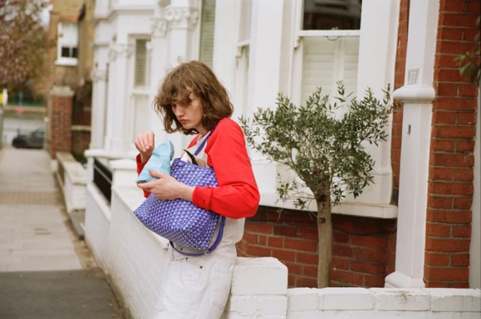 Model Katherine Vinten holding a purple patterned tote bag and snack pack by a residential townhouse wall.