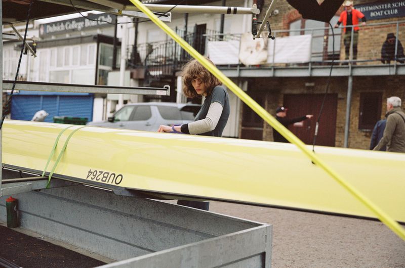 Model Katherine Vinten standing behind a bright yellow rowing boat at a riverside boat club for Moynat.
