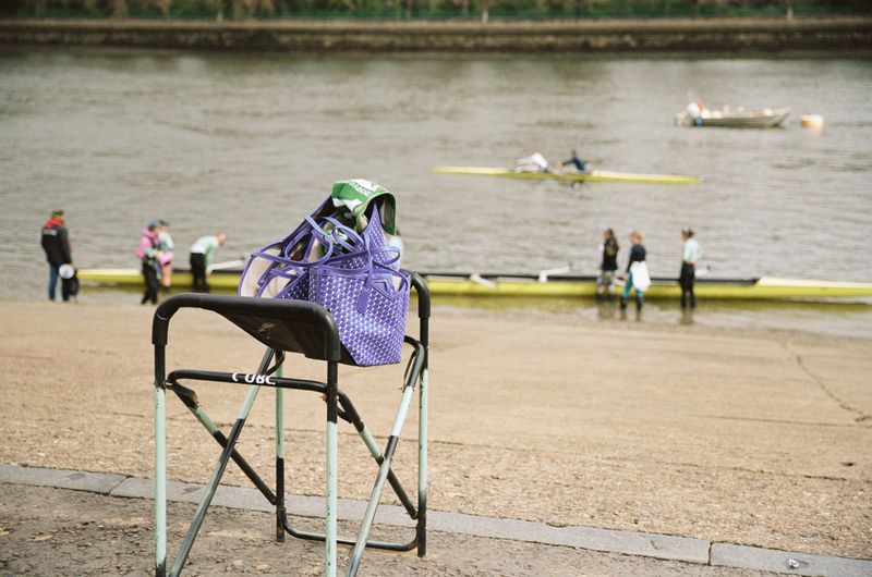 Purple monogram pattern tote bags resting on a folding rowing rack by a riverbank with rowing shells in the background.