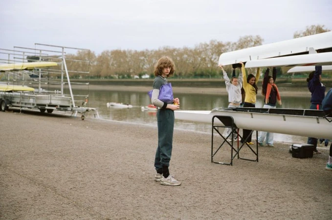 Model Katherine Vinten holding a purple patterned tote bag while standing near rowing shells on a riverbank.