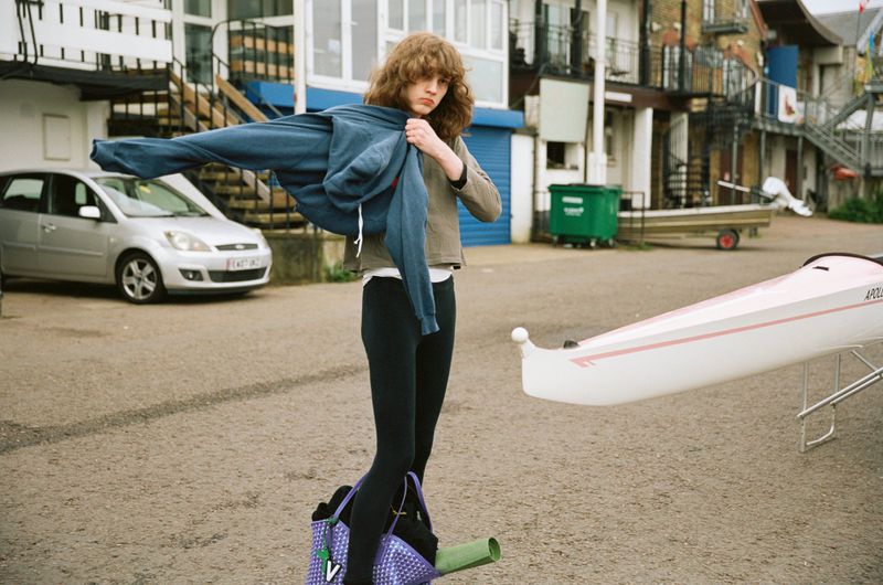 Model Katherine Vinten standing outdoors in black leggings and putting on a blue hoodie next to a purple patterned Moynat tote bag.
