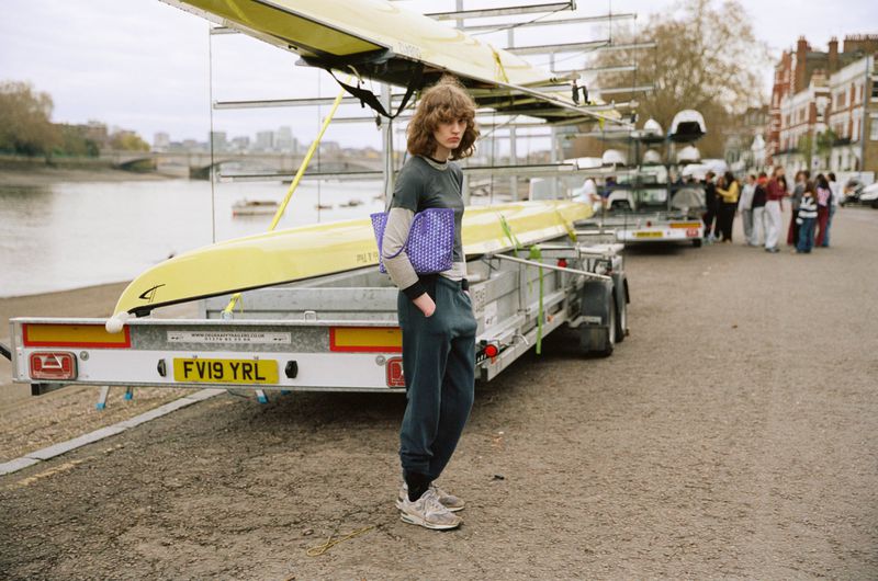 Model Katherine Vinten holding a purple Moynat tote bag beside a yellow rowing boat trailer near a riverbank.
