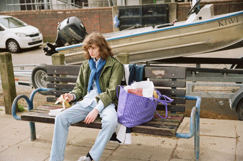 Model Katherine Vinten sits on a wooden bench holding a banana next to a purple patterned Moynat tote bag.