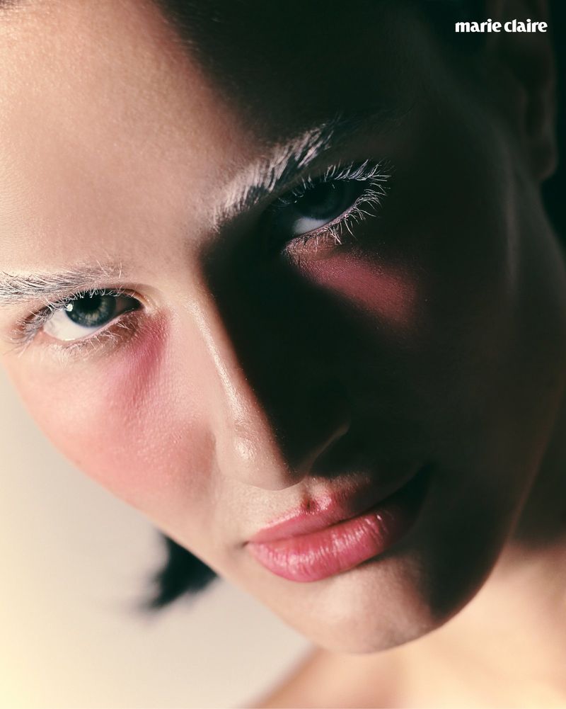 Close-up beauty portrait of a woman with white mascara, white eyebrows, and pink blush looking into the camera with dramatic shadow.