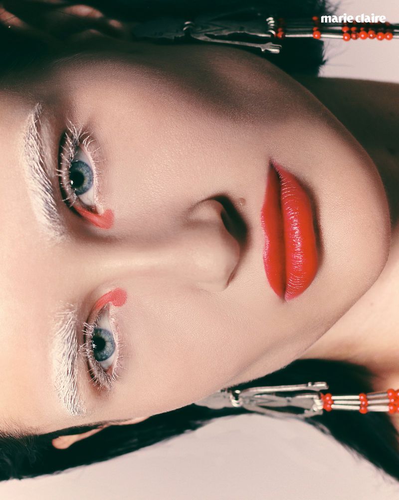 Close-up beauty portrait of a woman with bleached white eyelashes, white eyebrows, and bright red lipstick wearing beaded earrings.