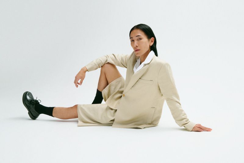A person with long dark hair is sitting on the floor in a beige suit with black shoes in a fashion style studio portrait.