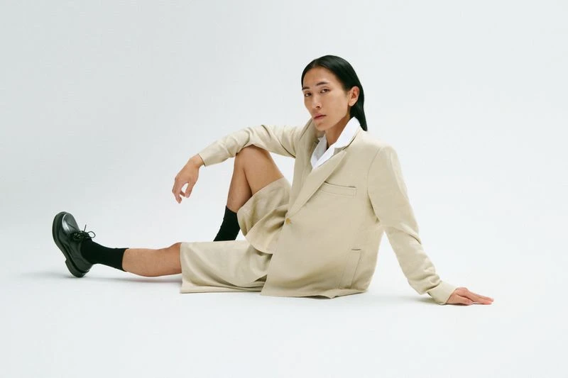 A person with long dark hair is sitting on the floor in a beige suit with black shoes in a fashion style studio portrait.