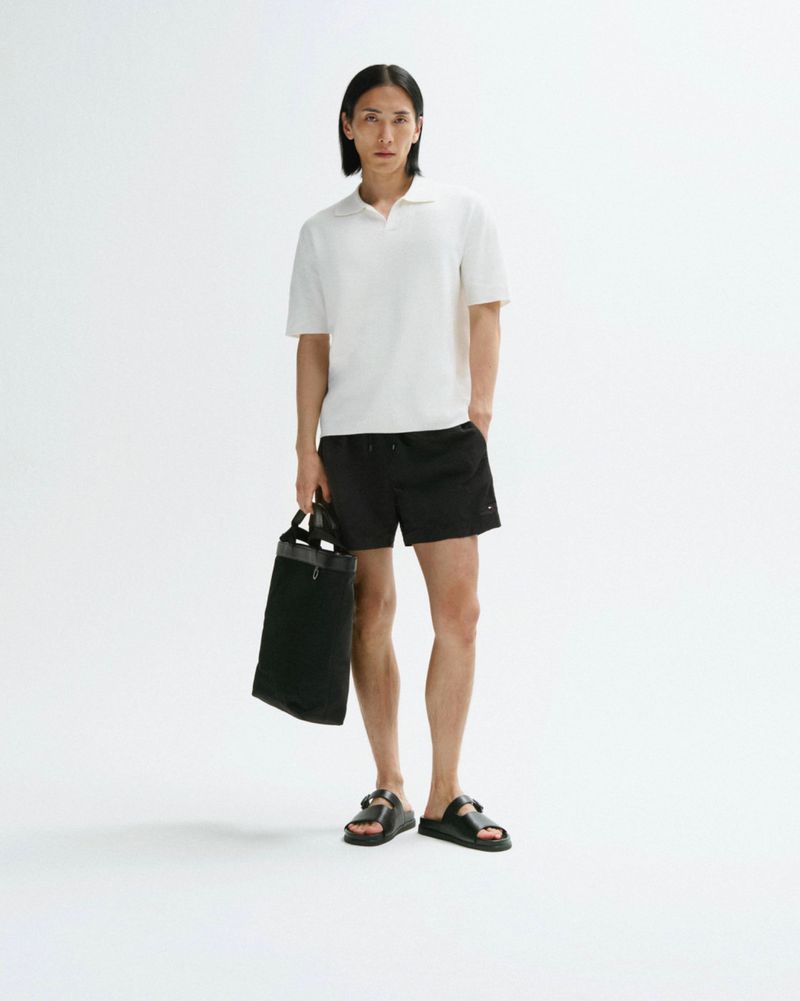 Full shot of a slender man in a polo shirt and shorts, holding a tote bag against a white backdrop, showcasing casual fashion.