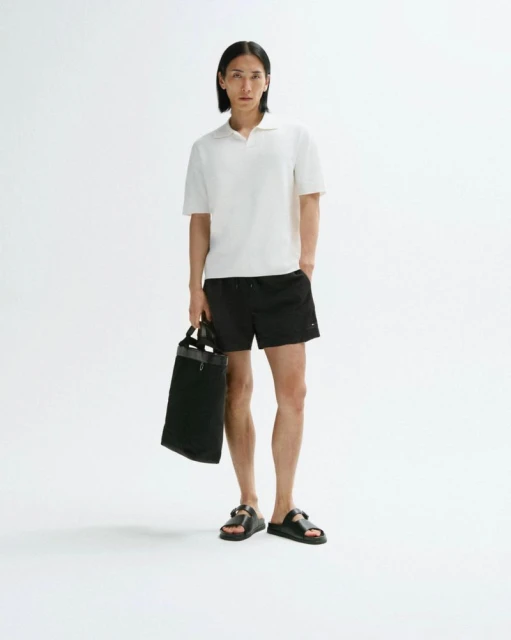 Full shot of a slender man in a polo shirt and shorts, holding a tote bag against a white backdrop, showcasing casual fashion.