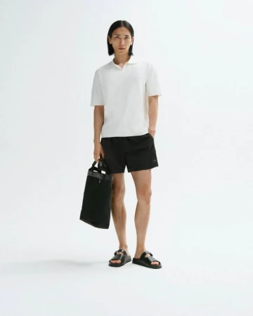 Full shot of a slender man in a polo shirt and shorts, holding a tote bag against a white backdrop, showcasing casual fashion.