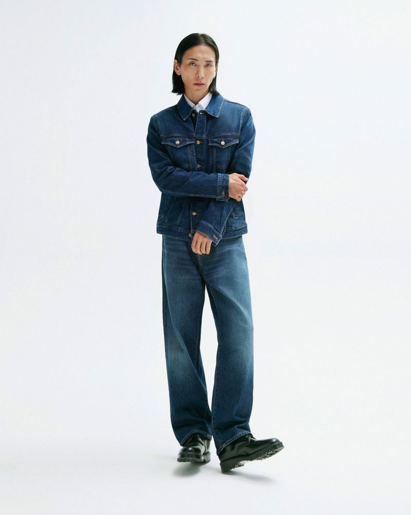 Full-length shot of an adult male with black hair wearing a dark blue denim jacket and jeans in a white studio.