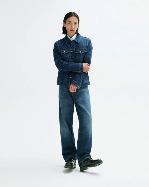 Full-length shot of an adult male with black hair wearing a dark blue denim jacket and jeans in a white studio.