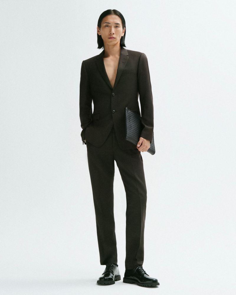 An Asian male fashion model wears a brown suit and black patent leather dress shoes against a white backdrop in a studio shot.