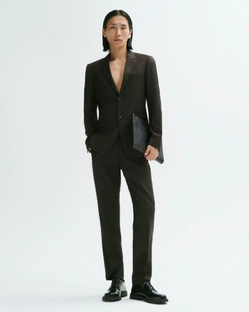 An Asian male fashion model wears a brown suit and black patent leather dress shoes against a white backdrop in a studio shot.