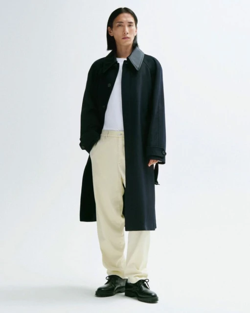 Asian male model poses in a navy coat, white t-shirt, and light-colored pants against a white backdrop in a fashion forward style.