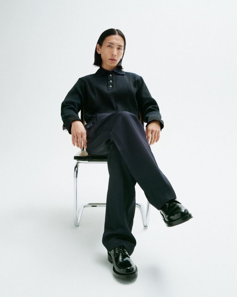 An adult male sits cross-legged on a chrome chair wearing a navy polo shirt, navy pants, and black leather shoes in a photo studio.