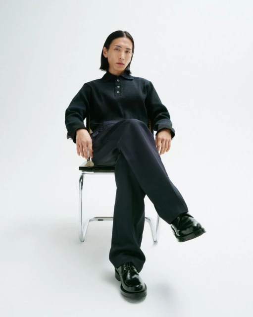 An adult male sits cross-legged on a chrome chair wearing a navy polo shirt, navy pants, and black leather shoes in a photo studio.