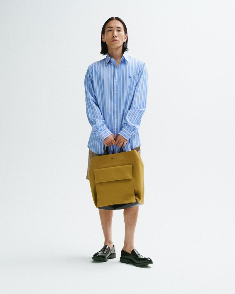 Full-length studio shot of an adult male model in a striped shirt, carrying a mustard-colored tote bag, posing against a white backdrop.