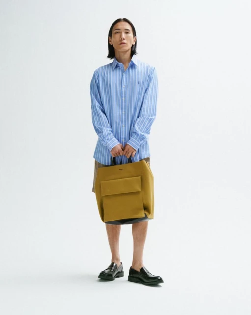 Full-length studio shot of an adult male model in a striped shirt, carrying a mustard-colored tote bag, posing against a white backdrop.